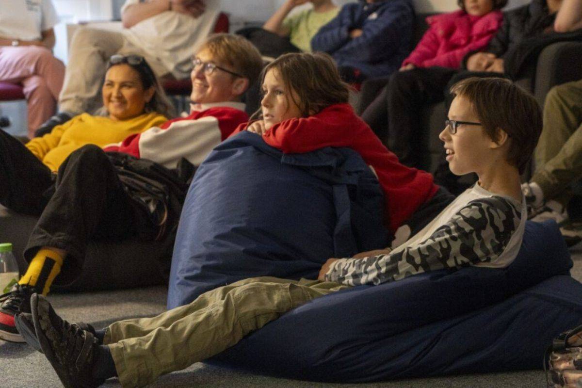 A group of children and young adults sit on beanbags and chairs, watching someone.